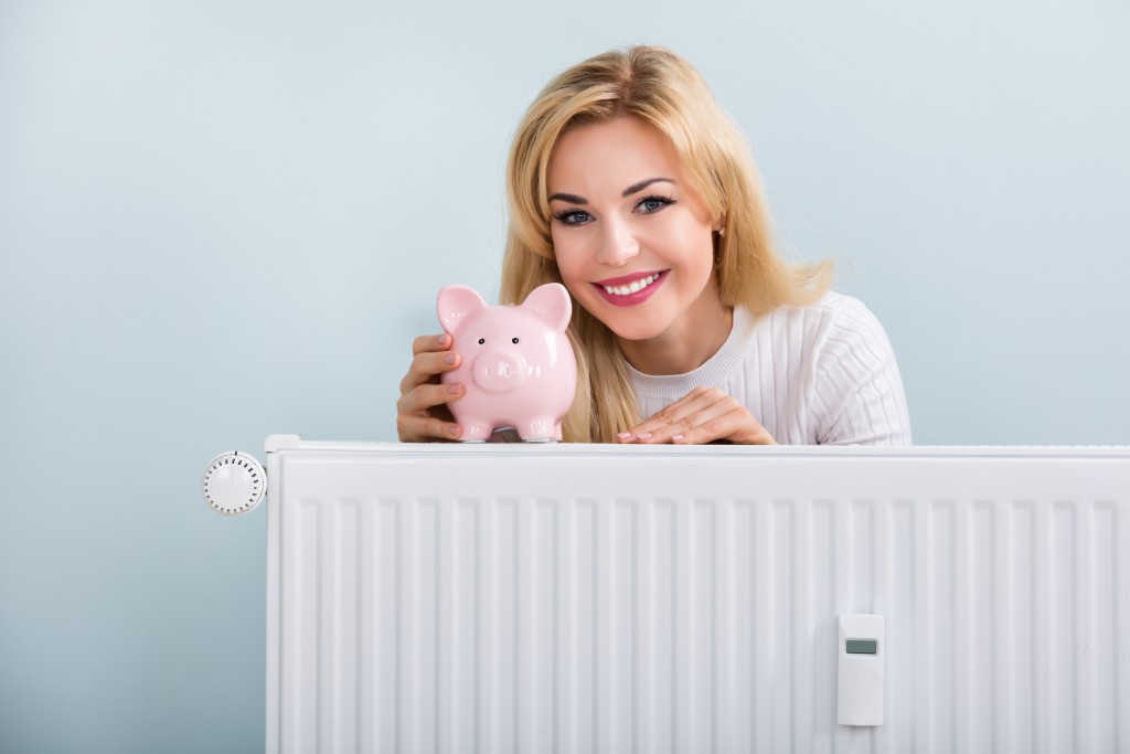 Young Happy Woman With Piggybank On Radiator At Home