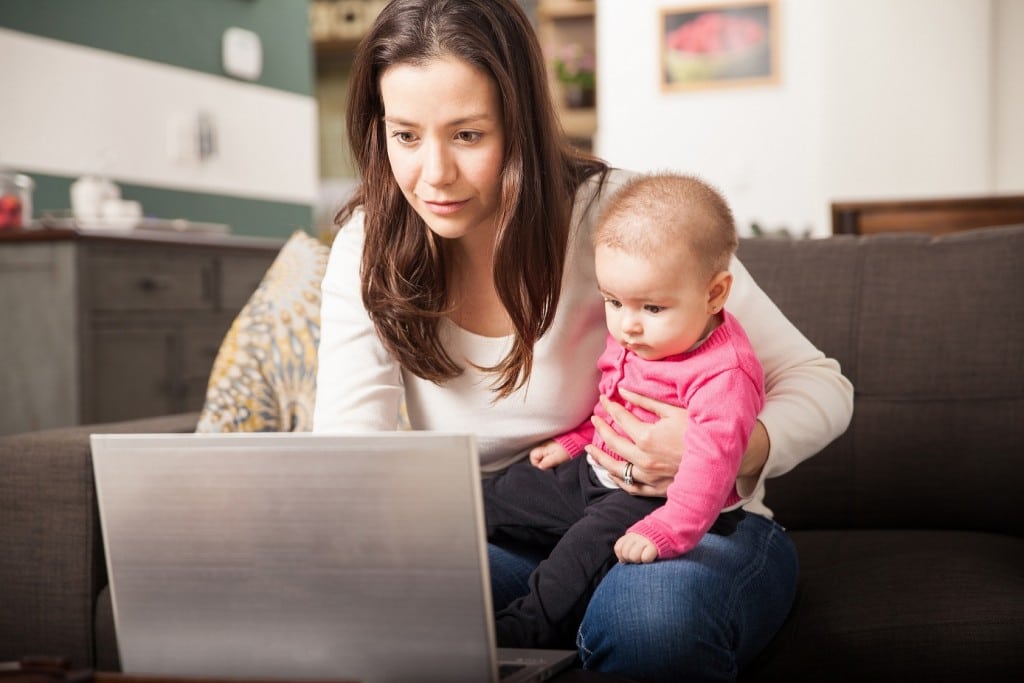 Pretty young single mom working at home on a laptop computer while holding her baby girl